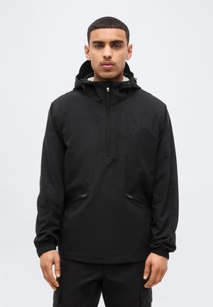 Jeune homme portant un anorak à capuche noir avec des poches zippées et des poignets élastiques, debout devant un fond uni gris clair.