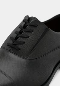 Chaussure en cuir noir avec une surface texturée, comprenant six paires d'œillets et un bout arrondi. Couture lisse le long du bord de la semelle.