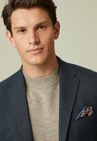 Blazer blu scuro su un maglione a coste beige chiaro, con un fazzoletto da taschino a fantasia con accenti blu, rosa e verde.