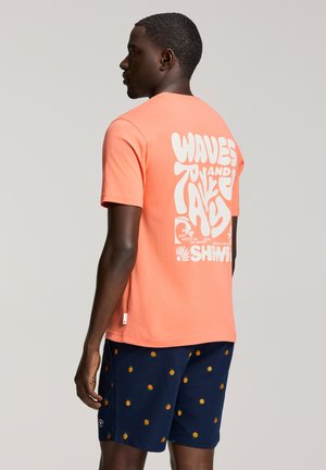 Mężczyzna w koralowym T-shircie z białym nadrukiem graficznym i granatowych szortach w drobny wzór pomarańczowych owoców, stojący bokiem na jasnym tle.