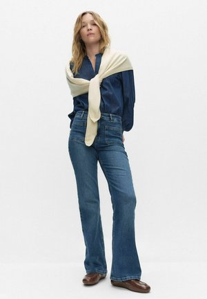 Denimskjorte i mørkeblå med knapper foran, kombineret med lyseblå vidde jeans og en cremefarvet striktrøje bundet ved skuldrene. Brune slip-on sko.