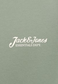 Tejido verde con textura suave, con el texto blanco "Jack & Jones" y "ESSENTIALS DEPT." impreso de manera prominente.