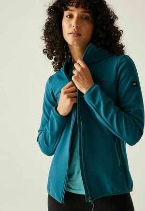 Mujer con cabello rizado cerrando la cremallera de una chaqueta softshell color teal, vistiendo una camiseta teal y pantalones negros, sobre un fondo claro y liso.