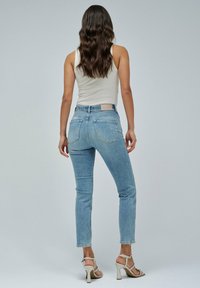 Vaqueros de tiro alto azul claro con un corte ajustado, que cuentan con bolsillos traseros cosidos y un parche de cuero marrón en la cintura. Combinados con sandalias beige.