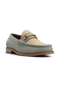 Mocasín verde de cuero con parte superior de mimbre beige, que presenta un acento en tono dorado y un diseño texturizado. Suela de madera marrón con perfil minimalista.