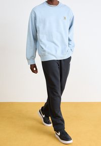 Ljusblå sweatshirt gjord av mjukt tyg, med ribbad mudd och en liten logotyp-patch. I kombination med mörkgrå träningsoverallbyxor och svarta sneakers.