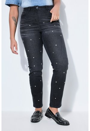 Persona vistiendo jeans negros con pequeños remaches plateados y mocasines negros con tachuelas, de pie contra un fondo blanco.