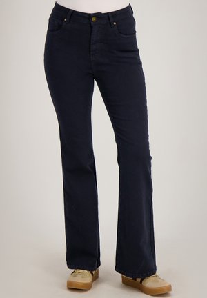 Vrouw draagt donkerblauwe bootcut jeans en beige sneakers, staand tegen een neutrale achtergrond.