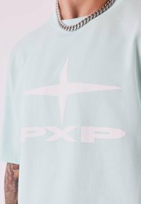 Camiseta de algodón de color azul claro con un logotipo estilizado en blanco que incluye las letras "PXP". El diseño presenta un acento gráfico de estrella por encima del texto.