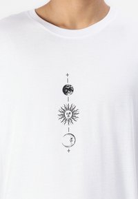 T-shirt bianco in cotone con una linea verticale di illustrazioni grafiche nere: Terra, sole con raggi e luna crescente, accompagnati da piccole croci.
