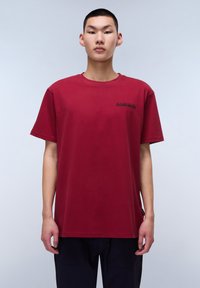 Camiseta de algodón color burdeos con mangas cortas, cuello redondo y un pequeño logotipo negro en el lado izquierdo del pecho. Corte recto y textura suave.