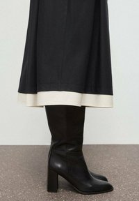Bottes en cuir noires montantes jusqu'au genou avec un talon carré et un bout pointu, associées à une jupe noire et crème à contraste. Texture lisse et design simple.