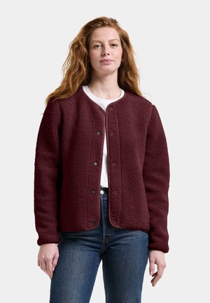 Kvinde med langt rødt hår iført en vinrød fleecejakke over en hvid top og blå jeans, stående mod en ensfarvet baggrund.