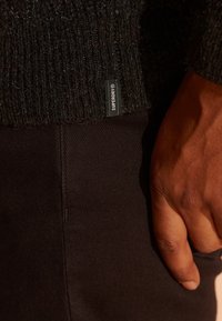 Zwarte gebreide trui met geribbelde textuur, voorzien van een klein "Superdry" label. Donkerbruine, slim-fit broek die gedeeltelijk zichtbaar is, met een gladde stof.
