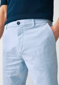 Helle blaue Stoffhose mit glatter Textur, featuring a button closure und seitlichen Taschen, teilweise über einem marineblauen Oberteil gezeigt.