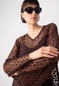 Brown lace top featuring a V-neck, long sleeves, and intricate floral patterns. Worn over a black underlayer. Black geometric sunglasses.