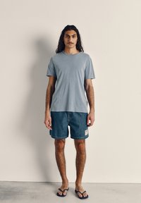 Light blue cotton T-shirt paired with dark denim shorts. Shorts feature a patch label; complementing blue flip-flops complete the outfit.