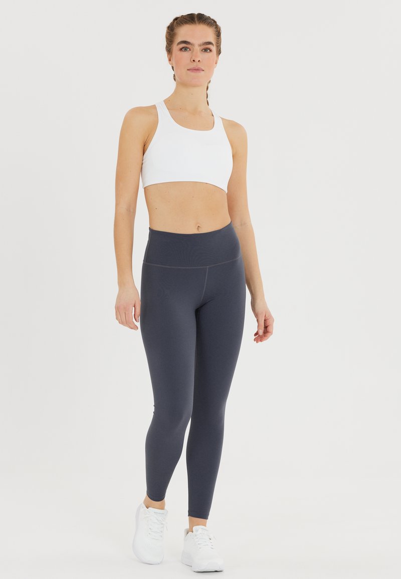 Valkoinen urheil Bra yhdistettynä korkeavyötäröisiin tummanharmaisiin leggingsseihin. Leggingsseissä on sileä pinta ja niissä on minimaaliset sauman yksityiskohdat. Valkoiset urheilu kengät.