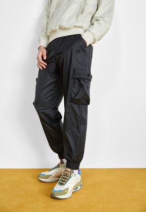 Person iført sorte Nike cargo-bukser med elastiske manchetter, en lysefarvet sweatshirt og beige sneakers med flere farver, der står på en karrygul gulv.