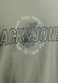 Zelená tkanina s velkým tmavým textem "JACK & JONES" a menším bílým kruhovým textem, který zní "Celosvětově Kreativní Proces v Pohybu".