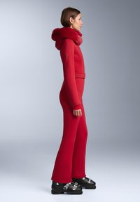 Piros síruha texturált felülettel és szőrmebéléssel ellátott kapucnival, testhezálló szabással, öves derékkal és bővülő szárakkal, fekete síbakancsokkal párosítva.