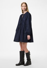 Vestido de denim azul marinho com gola redonda, mangas longas e cintura ajustada, apresentando uma fenda e detalhe de laço na parte da frente. Botas pretas altas até o joelho com fivelas.