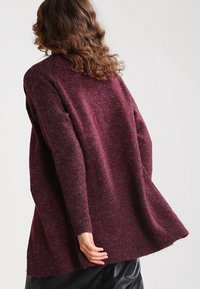 Bordeauxfärgad stickad kofta med en lös passform, långa ärmar och en strukturerad yta. Har rundad nederkant och ribbade ärmar.