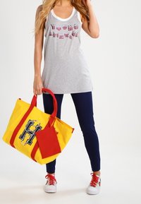 Szara koszulka bez rękawów z białym dekoltem, niebieskie legginsy, czerwone trampki oraz duża żółta torba na ramię z czerwonymi paskami i grafiką litery "H".