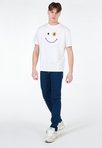 T-shirt bianca in cotone con faccina sorridente nera e accento arancione. Abbinata a jeans blu e scarpe da ginnastica chiare, modello che cammina su uno sfondo neutro.