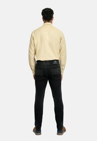 Jeans negros con costuras amarillas, diseño ajustado y dos bolsillos traseros. Combinados con una camisa de manga larga color beige claro y zapatos marrones.
