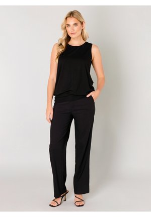 Blonde vrouw die een mouwloze zwarte top, zwarte wijde broek en zwarte sandalen met hakken en bandjes draagt, staand met één hand in haar zak.