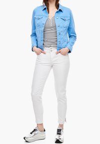 Hellblaues Denim-Jackett über einem gestreiften Oberteil, kombiniert mit weißen Hosen und silbernen Sneakers. Besitzt Knopfakzente und ein tailliertes Design.