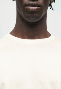 Gros plan sur la partie inférieure du visage d'une personne, montrant les lèvres, le cou et une partie d'une chemise couleur crème avec des dreadlocks courtes visibles sur un côté.