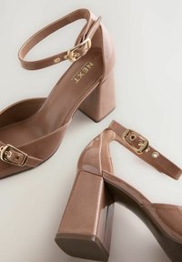 Beige patent leather high-heel sandals with a square heel, ankle strap, and gold buckle detail. Smooth texture with a glossy finish.