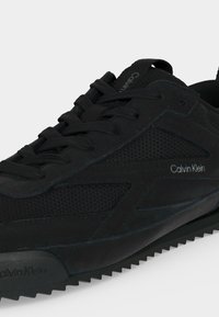 Baskets Calvin Klein noires avec panneaux texturés, lacets à l'avant et logo sur le côté et la languette, montrées en gros plan.