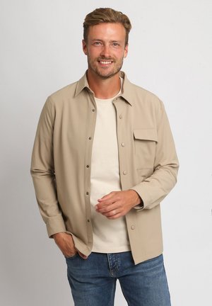 Lachende man met lichtbruin haar, beige overhemd met knopen over wit T-shirt en blauwe spijkerbroek, staand tegen een effen lichte achtergrond.