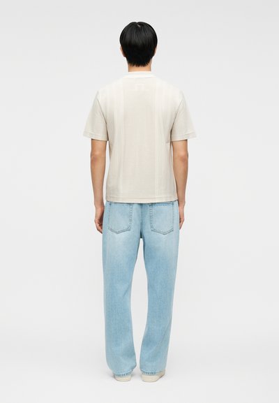 Άνδρας που φοράει κοντομάνικο μπεζ πουκάμισο με υφή και ανοιχτόχρωμα, φαρδιά τζιν, στέκεται γυρισμένος πλάτη πάνω σε μονόχρωμο φόντο.