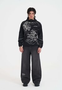 Felpa nera con design di dragone e nuvole bianche, con maniche raglan e tasca frontale. Indossata con jeans grigi larghi e calzature nere.