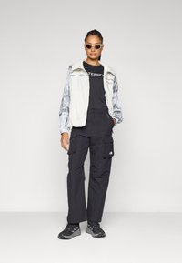 Light grey zip-up vest with marbled sleeves, black t-shirt, black cargo pants, and dark sneakers. The person wears light sunglasses.
