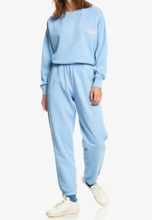 Vrouw draagt lichtblauwe sweater en bijpassende joggingbroek met witte sneakers, staande met één voet licht opgetild.