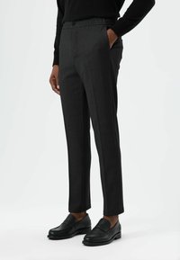 Donkergrijze, op maat gemaakte broek met een ontspannen tailleband, voorzien van zijzakken en een gladde stoftextuur, gecombineerd met zwarte loafers.