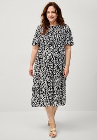 Schwarzes und weißes Blumenmuster-Kleid mit kurzen Ärmeln, gerafftem Ausschnitt, fließendem Stoff und gestuftem Saum. Getragen mit nudefarbenen Riemchensandalen.