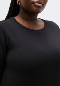 Κοντινό πλάνο ενός ατόμου που φορά μαύρη μπλούζα με στρογγυλή λαιμόκοψη και χρυσό σκουλαρίκι κρίκο, με πλεκτά μαλλιά σε ουδέτερο φόντο.