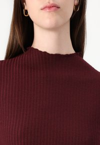 Burgunderfärgad ribbad topp med en texturerad tyg och scallopad halsringning, tillsammans med guldiga hoopsörhängen.