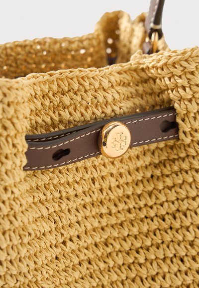 Κοντινή λήψη μπεζ υφαντής τσάντας με καφέ δερμάτινη επένδυση και χρυσό κουμπί με ανάγλυφο συμμετρικό λογότυπο.