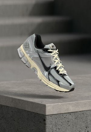 Γκρι και μαύρο τρέχον παπούτσι Nike με μπεζ κορδόνια που αιωρείται πάνω από μια γκρι τσιμεντένια άκρη και σκαλοπάτια στο φόντο.