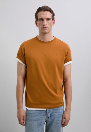 Mann mit kurzen braunen Haaren, der ein rostorangefarbenes Kurzarm-T-Shirt über einem weißen Hemd und hellblauen Jeans trägt, vor einem grauen Hintergrund stehend.