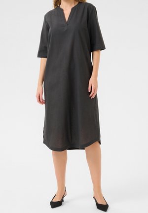 Kobieta ubrana w luźną czarną sukienkę midi z rękawami do łokcia i dekoltem w serek, zestawioną z czarnymi butami na obcasie typu slingback.