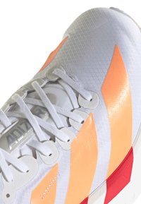 Bílé sportovní boty s oranžovými akcenty, texturovaný síťovaný svršek, ploché bílé tkaničky a červená gumová podrážka s bílými pruhy.