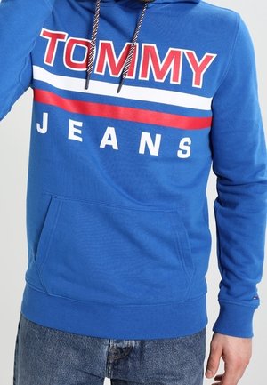 Niebieska bluza z kapturem z czerwono-białymi paskami i napisem „TOMMY JEANS”, noszona przez osobę w niebieskich dżinsach, widoczna ręka po boku.